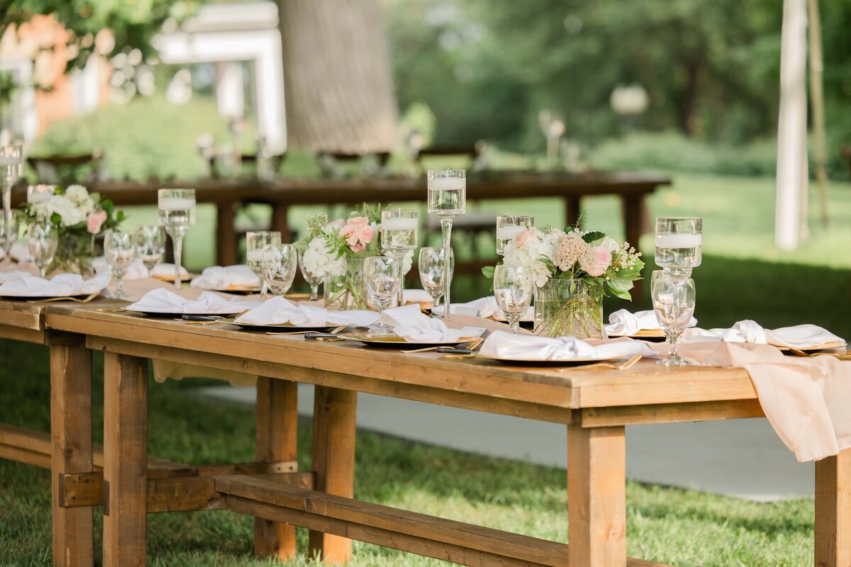 Elegant outdoor dining setup with wooden tables, floral centerpieces, and place settings in a lush, green park farm winery.