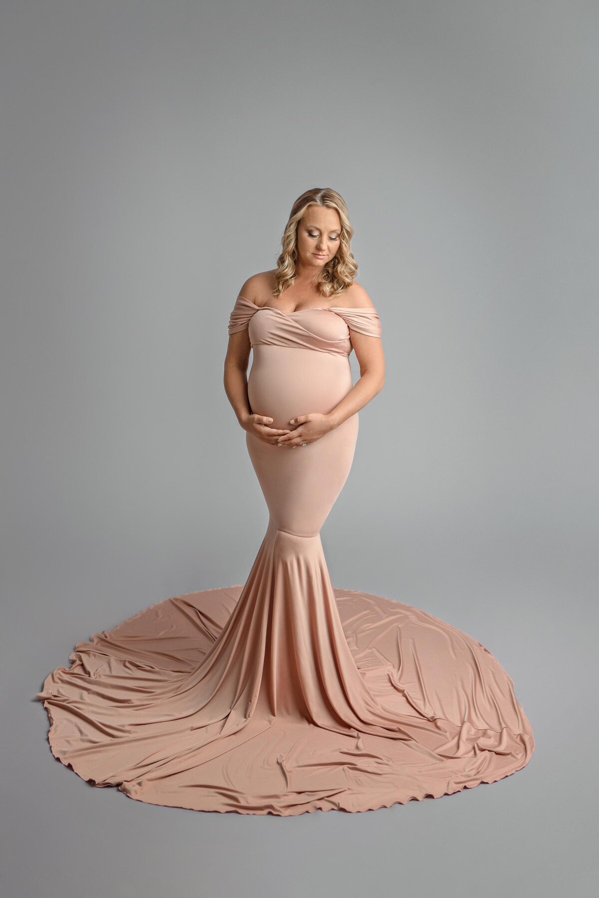 Expecting mom cradling belly dressed in pink flowing dress on grey backdrop