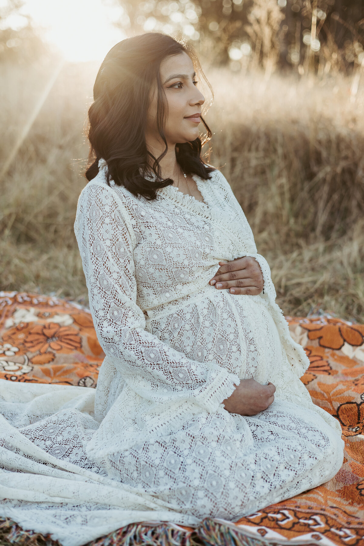 Pregnant woman sitting on her knees on orange picnic blanket while holding her baby bump