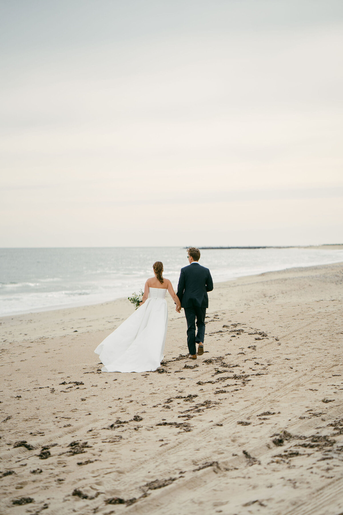 Couple holding hands walking on beach on wedding day, wedding planned by Unique Melody Events & Design (New England Wedding Planners)