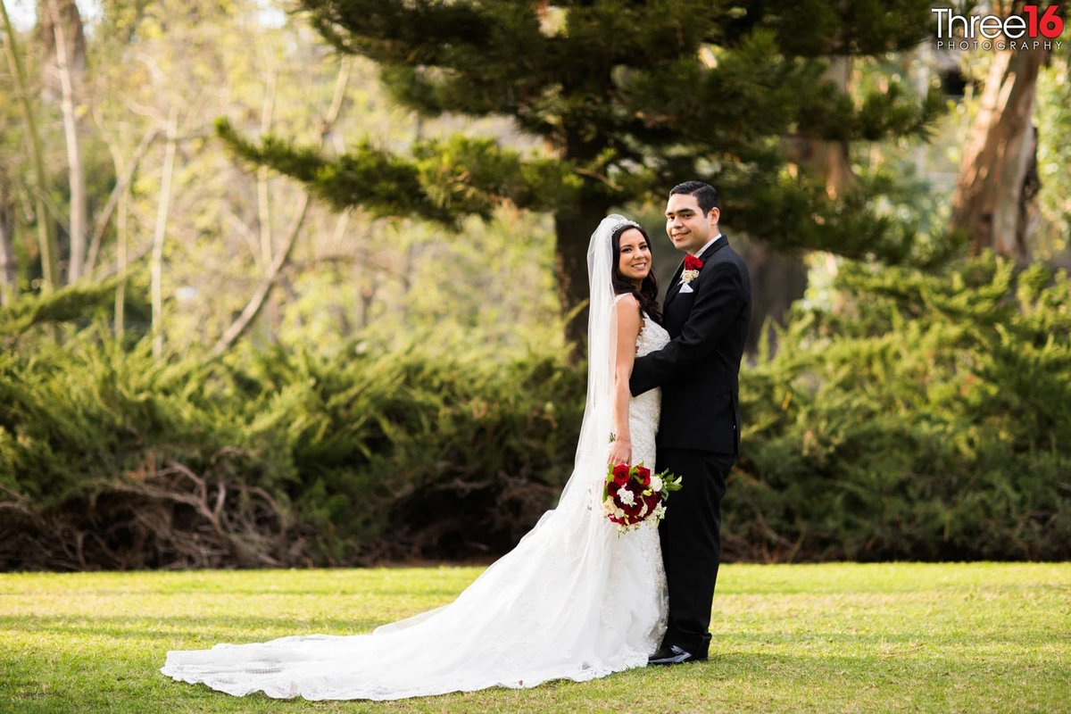Groom holds his Bride as they smile for the wedding photographer while standing in an open patch of green grass and green trees and bushes surround them