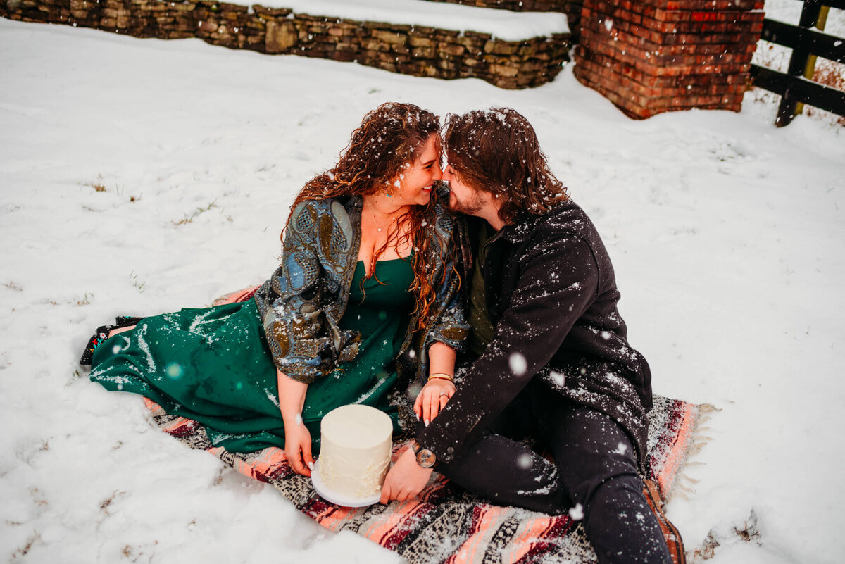 photo of a couple sitting on a blanket in the snow while holding a small white cake