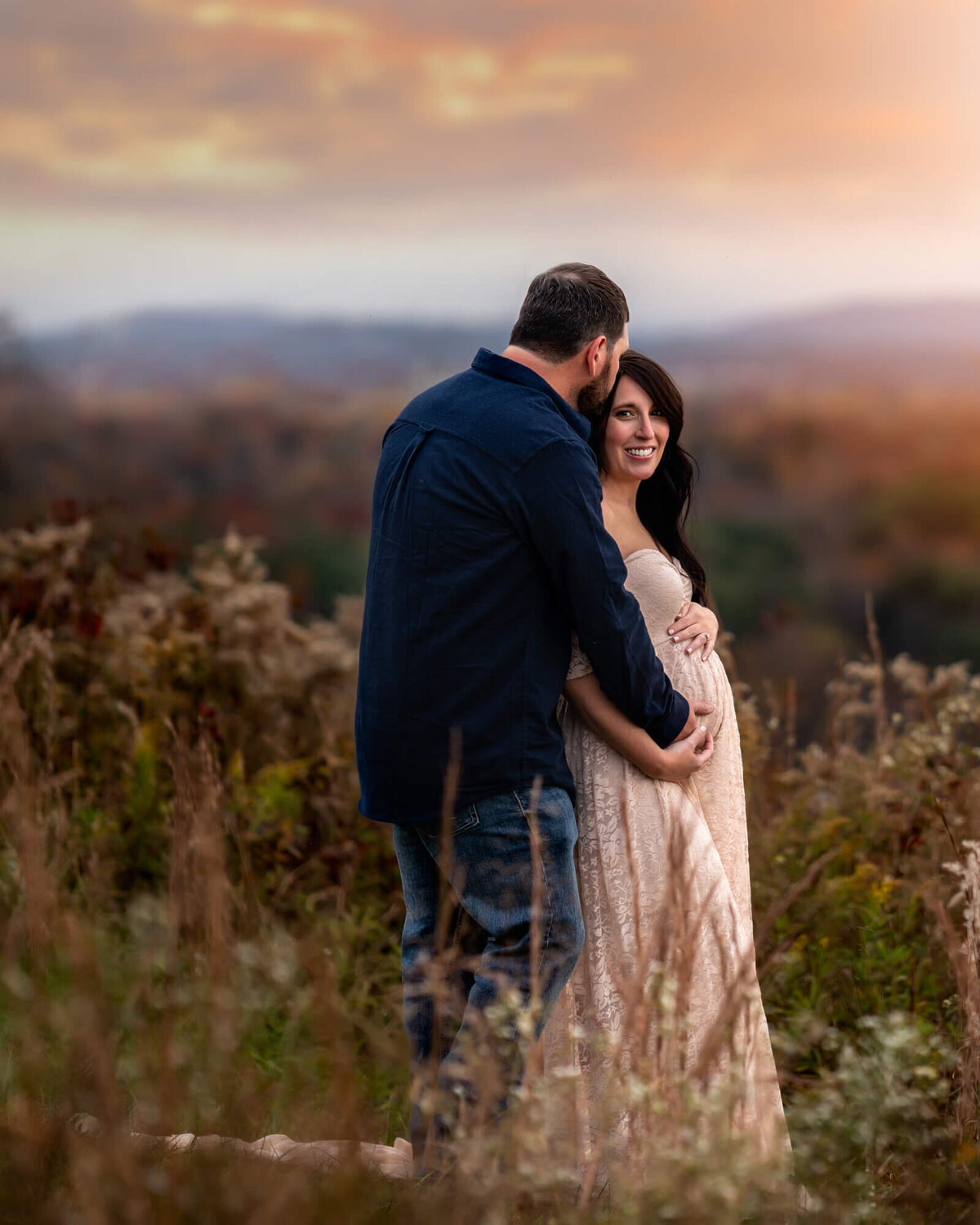 An expecting couple embrace while standing in the tall grass on a mountain top