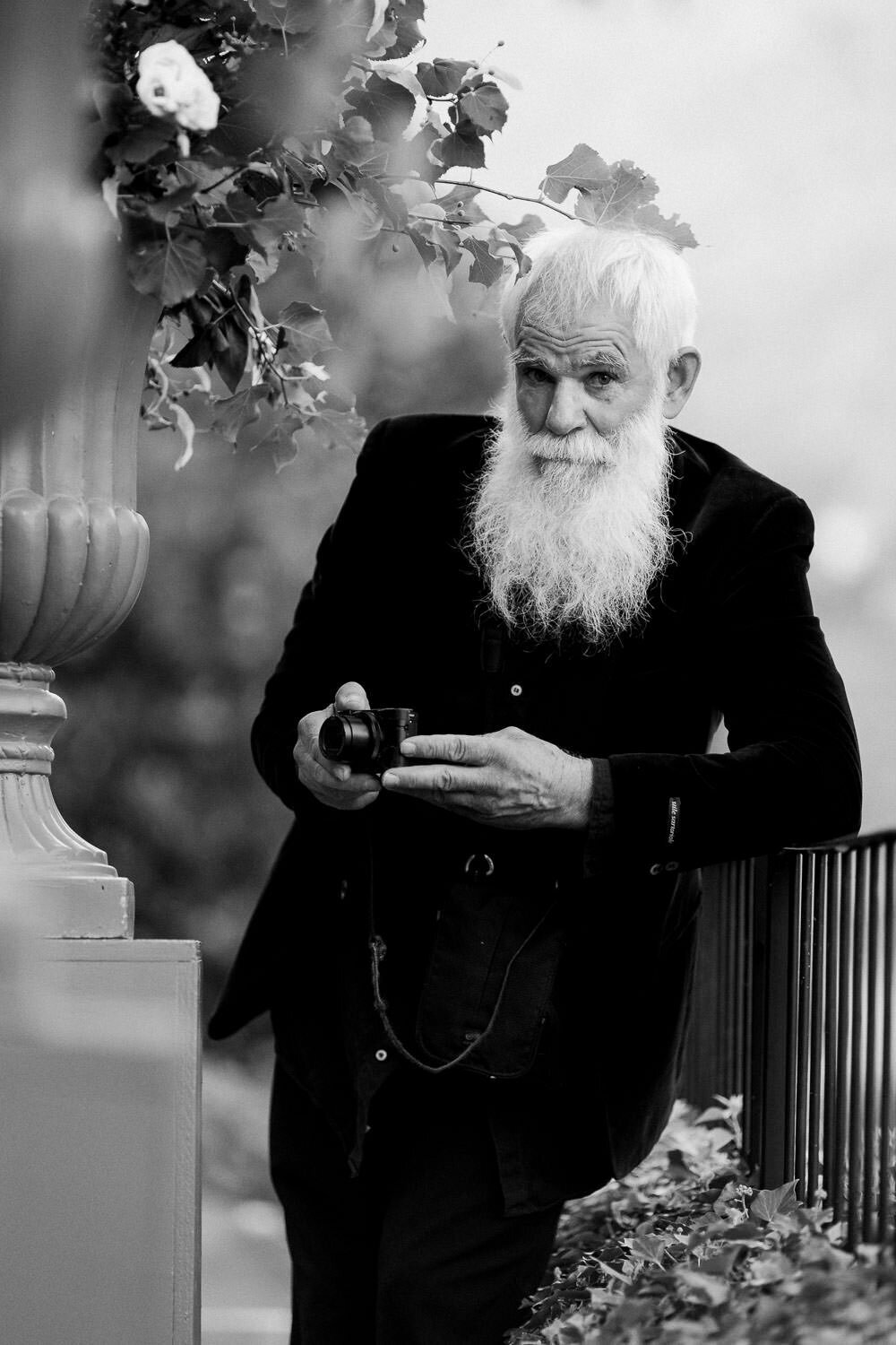 white bearded old man holding camera leaning against fence in black suit