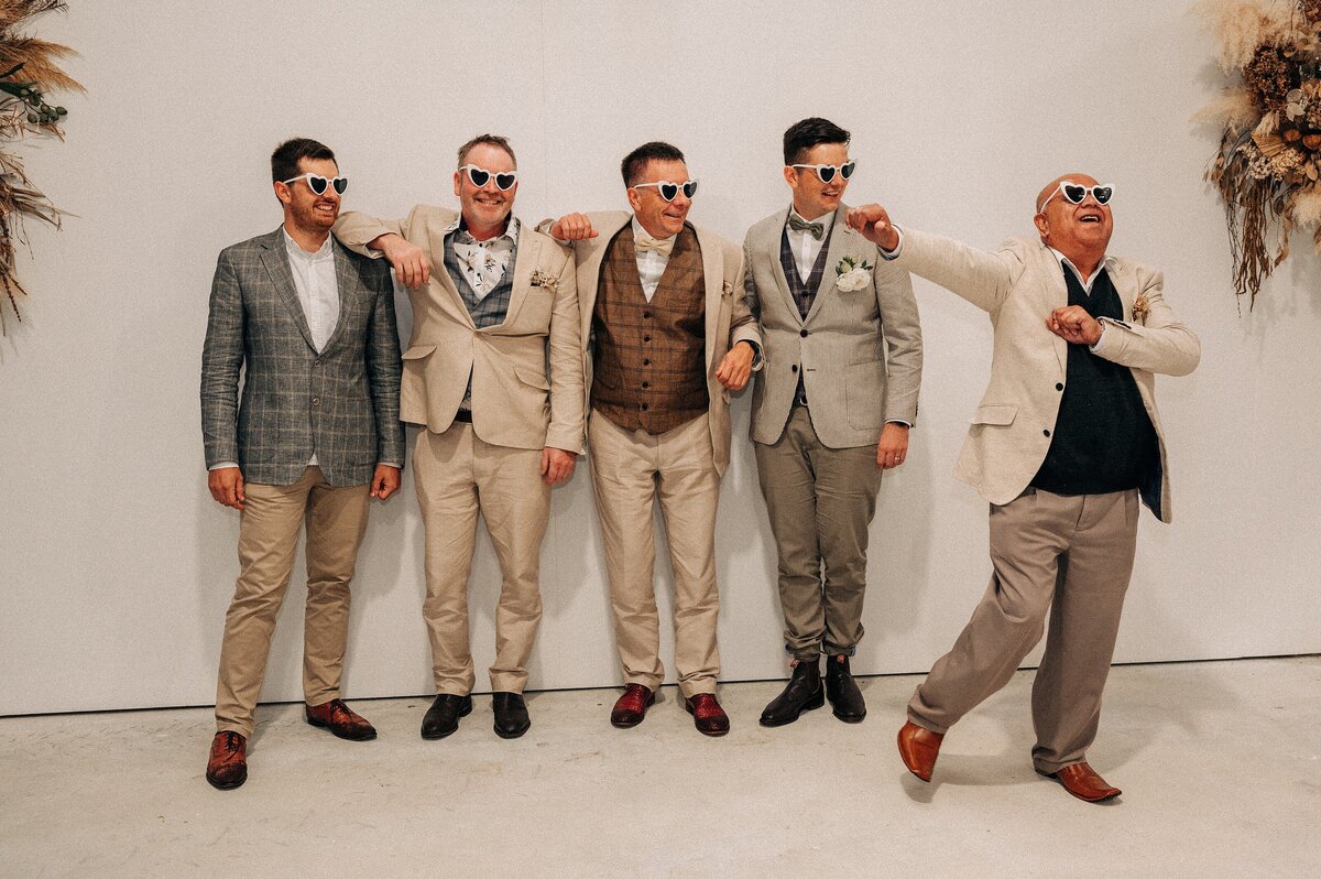 a groom and his family line up against a white wall wearing heart sunglasses while one man dabs