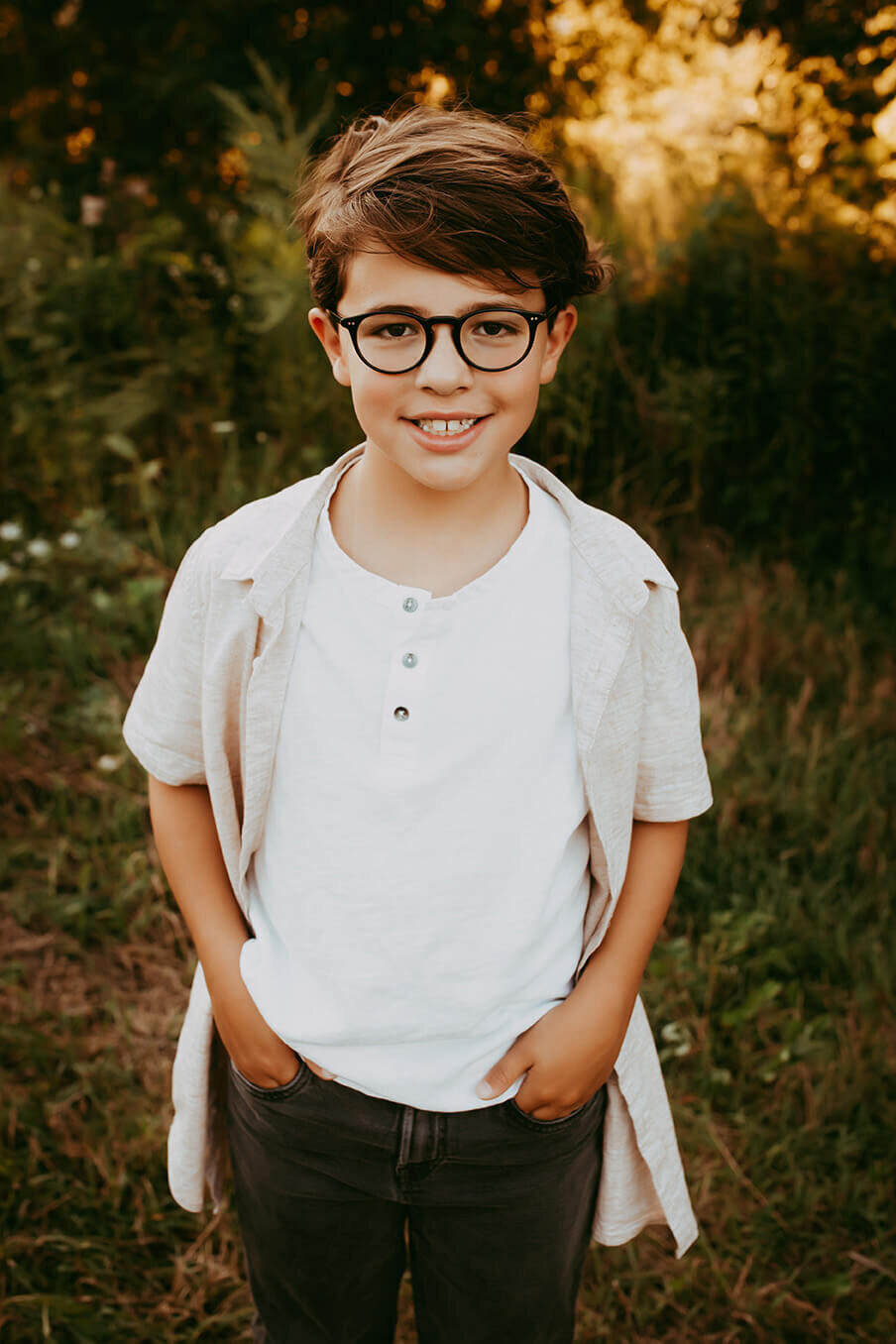an eight year old boy standing with his hands in his pockets  looking and smiling at the camera