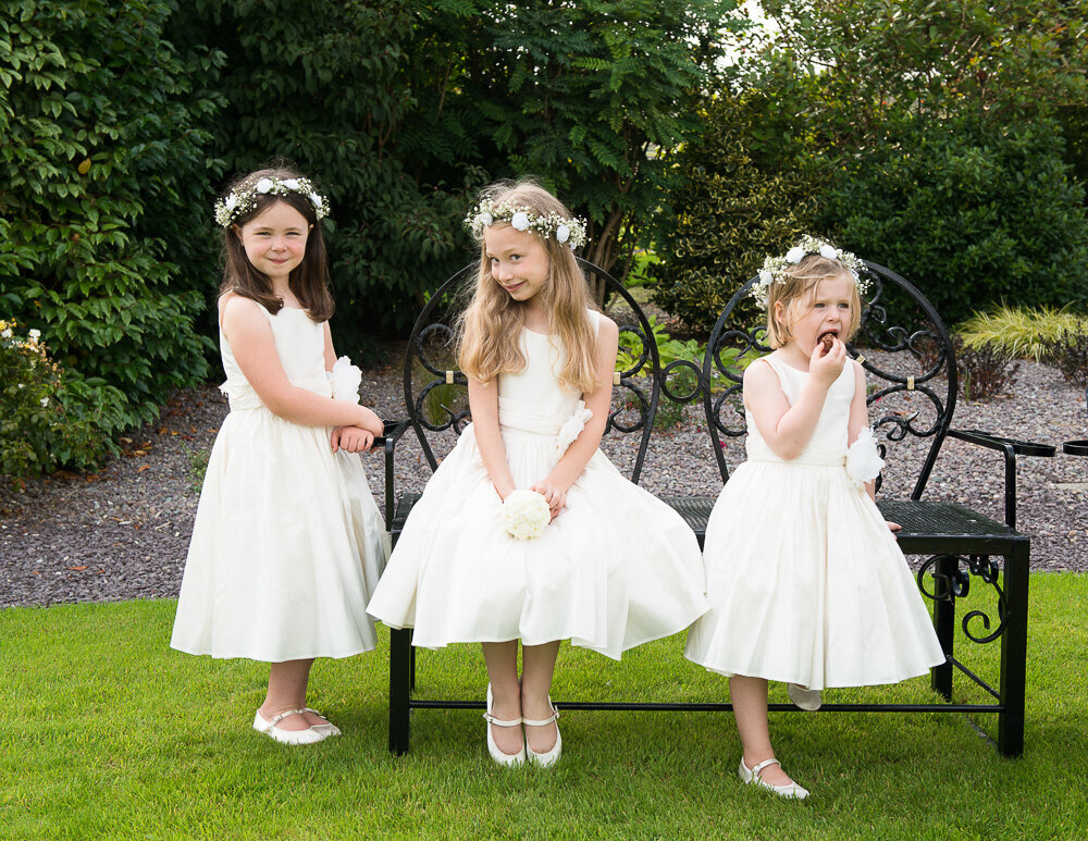 three flower girls sitting on a garden bench wearing white dresses and white flower head pieces