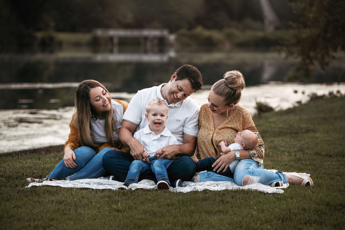 dad, mom holding an infant, teenage sister and  little brother laughing at each other while sitting on a blanket in the grass in from of a lake at sunset in the summer