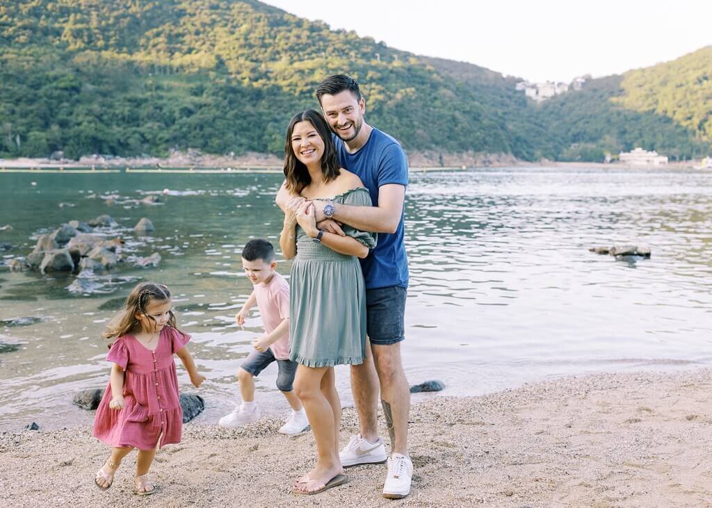 Daddy giving mommy a backhug while children play by the lakeside.