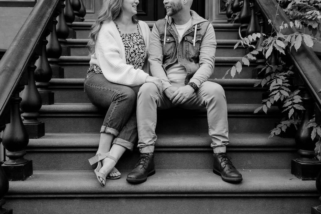 black and white photo of a woman with her arms on a man's leg as they sit on stairs and smile at each other