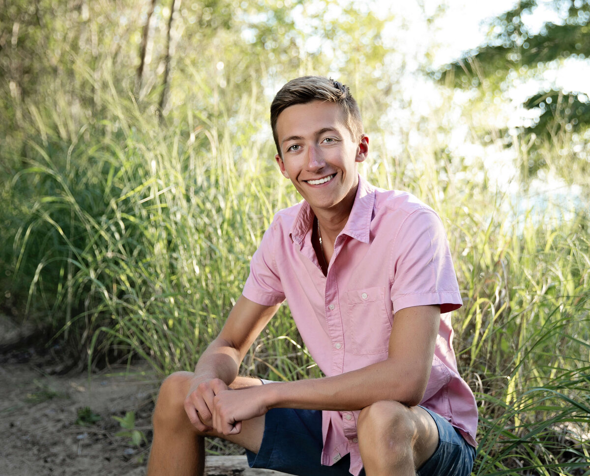 Senior portrait of a senior boy in a pink shirt sitting on a log at Presque Isle State Park
