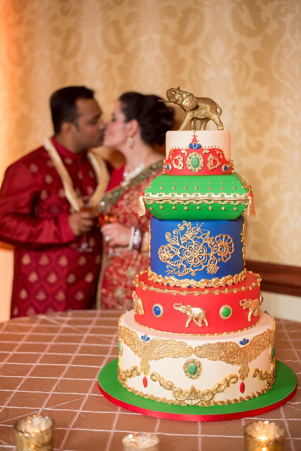 Indian bride and groom wearing red and gold kissing behind their green, blue and ivory wedding cake topped with a gold elephant and gold mandala decorations.