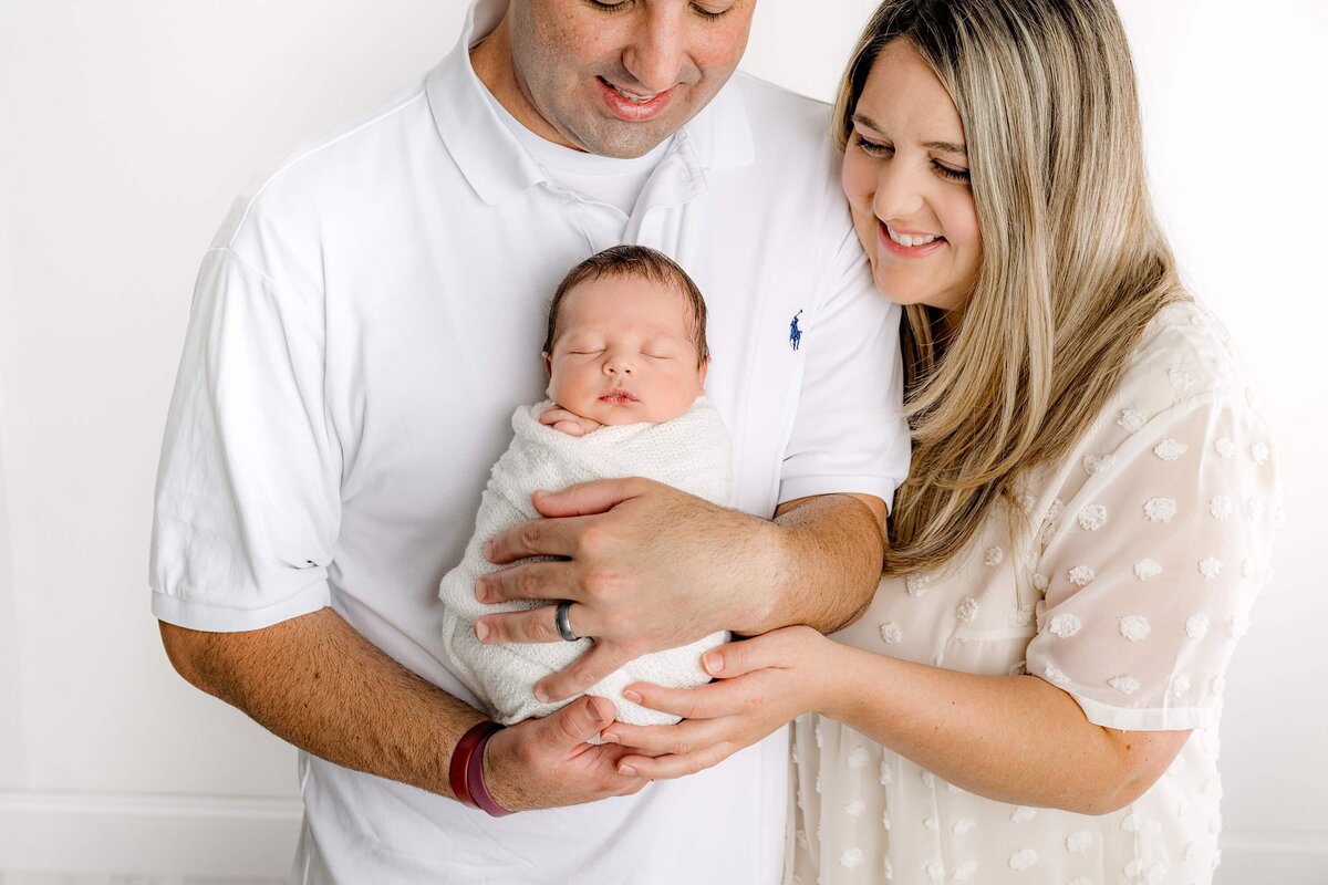 Father holds his newborn son wrapped and white with Mom standing beside him and her hand on the newborn baby