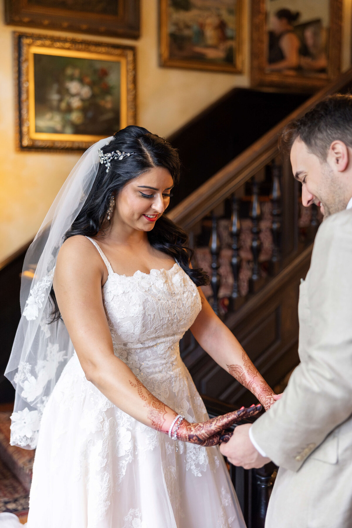 A bride in a white lace dress and veil, with henna on her hands, stands facing a groom in a beige suit. They hold hands in a warmly lit room with wooden stairs and framed artwork in the background.