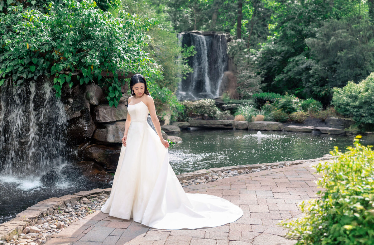 A bride in a white gown stands beside a flowing waterfall and lush greenery. The scene is serene, with sunlight filtering through the trees, casting a gentle light on the bride and the surrounding garden.