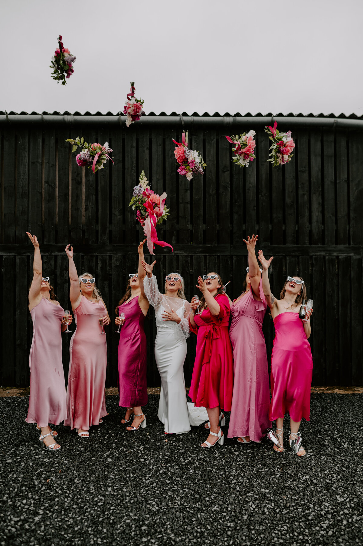 6 bridesmaids in pink bridesmaids dresses and a bride throw their pink bouquets in the air. They are all wearing white heart shaped sunglasses.