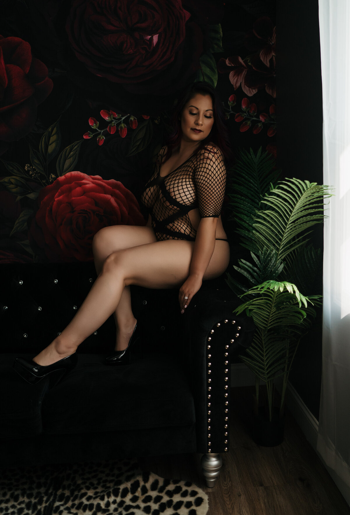 A woman in mesh lingerie gazes down her shoulder while sitting on the shoulder of a black couch in front of a floral tapestry