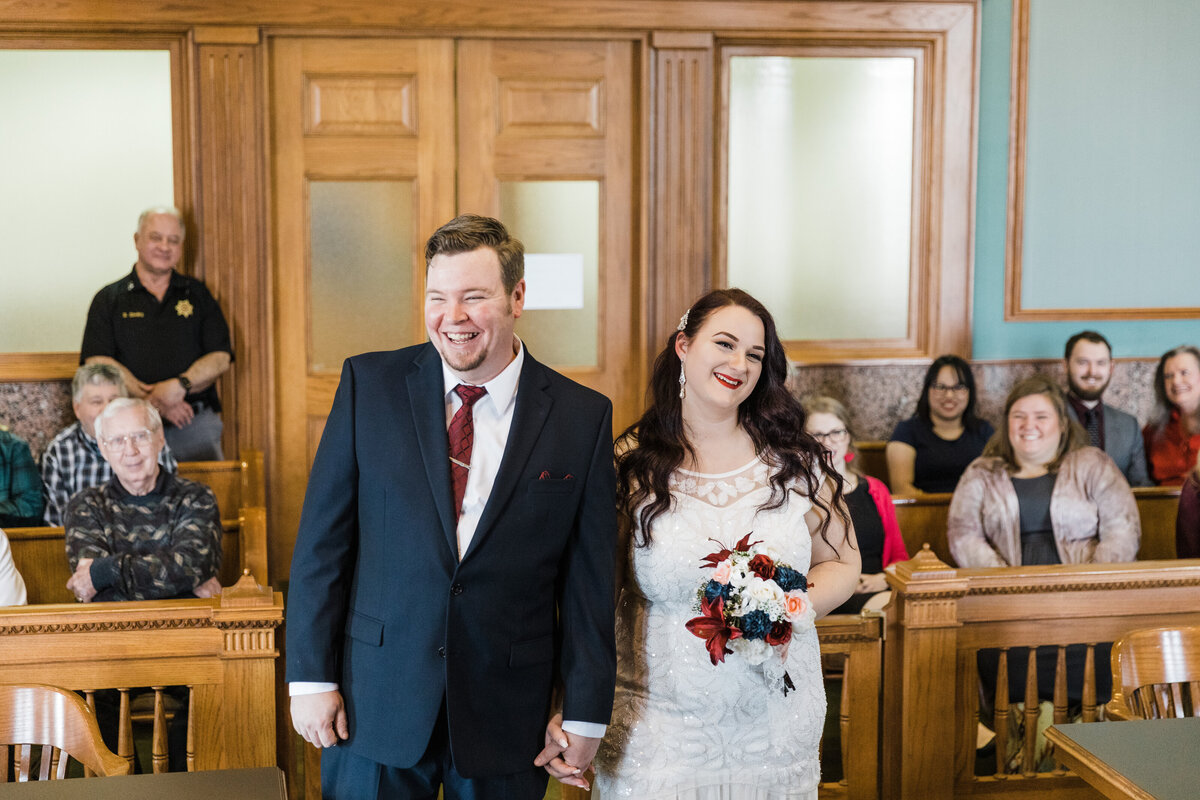 A candid shot of a bride and groom walking into the courtroom for their wedding ceremony at the Tarrant County Courthouse in Fort Worth, Texas. The bride is on the right and is wearing a detailed, white dress and is holding a bouquet. The groom is on the left and is wearing a blue suit with a red tie. They both are holding hands and smiling joyfully. Their friends and family can be seen seated in background.