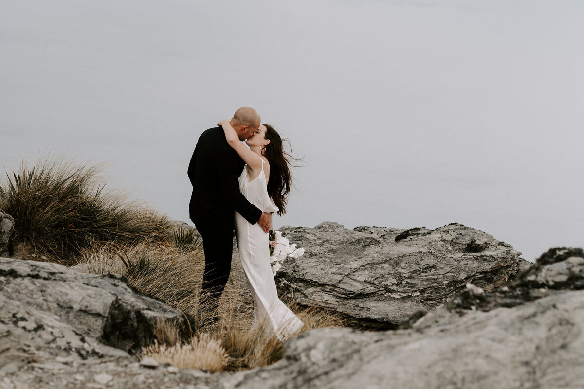 The Lovers Elopement Co - couple kiss on rocks top of mountain, heli wedding