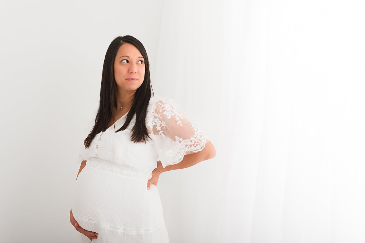 Expectant mother in a white dress holding her belly while looking off into the distance with a hopeful expression. Taken by Minneapolis Maternity Photographer, Fig and Olive Photography.