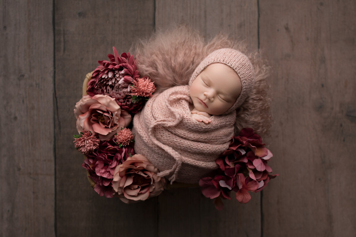 Fine art newborn photos captured by best Philadelphia newborn photographer Katie Marshall. Baby girl is sleeping swaddled in a dusty pink with a matching knit bonnet. Baby is sleeping atop of a fuzzy rug adorned with flowers.