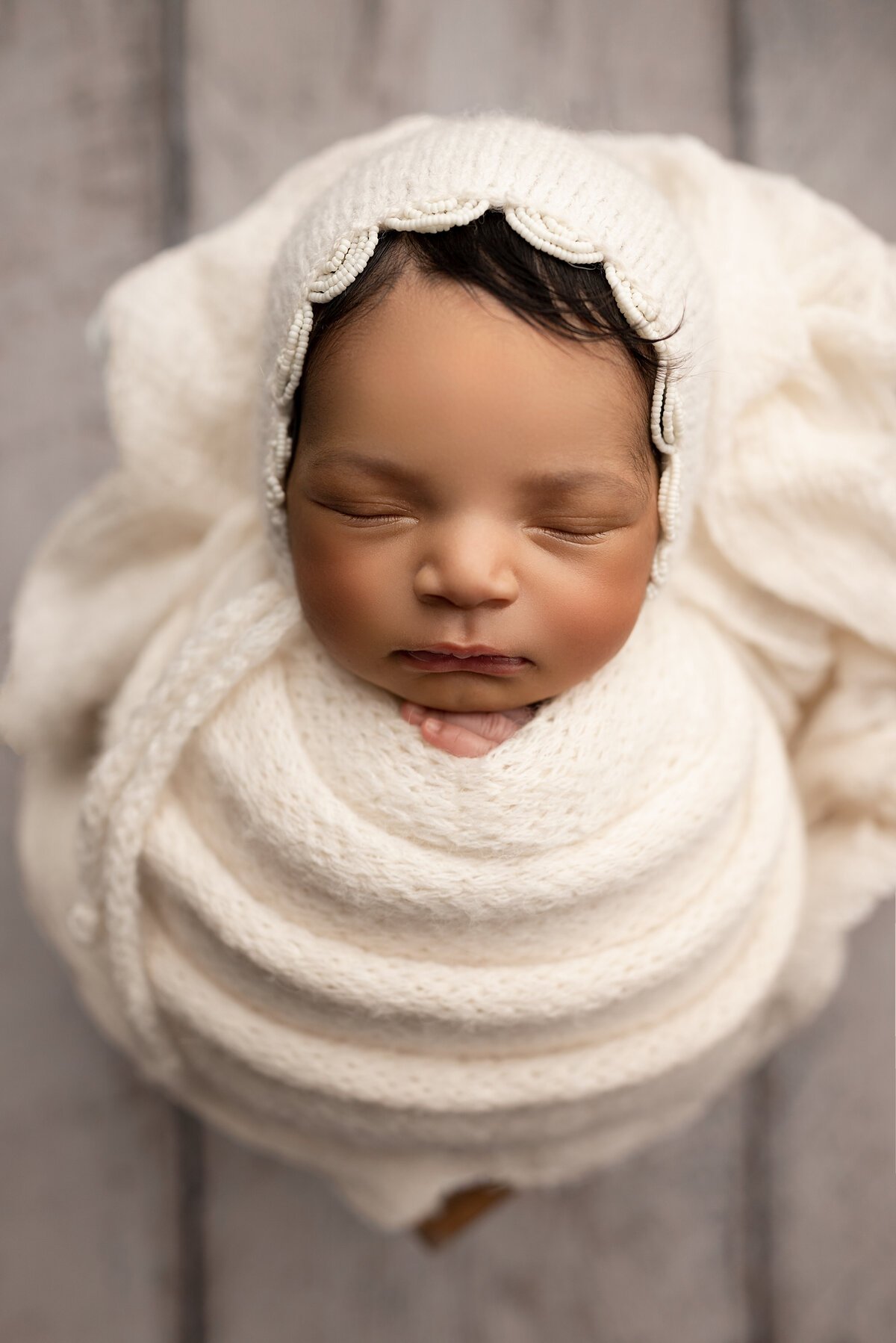 Fine art newborn photo captured by Philadelphia's best newborn photographer Katie Marshall. Baby swaddled in white with matching bonnet with scallop detail. Baby's fingers are peeking out of the top of the swaddle. Baby's long dark hair is peeking out of the bonnet.