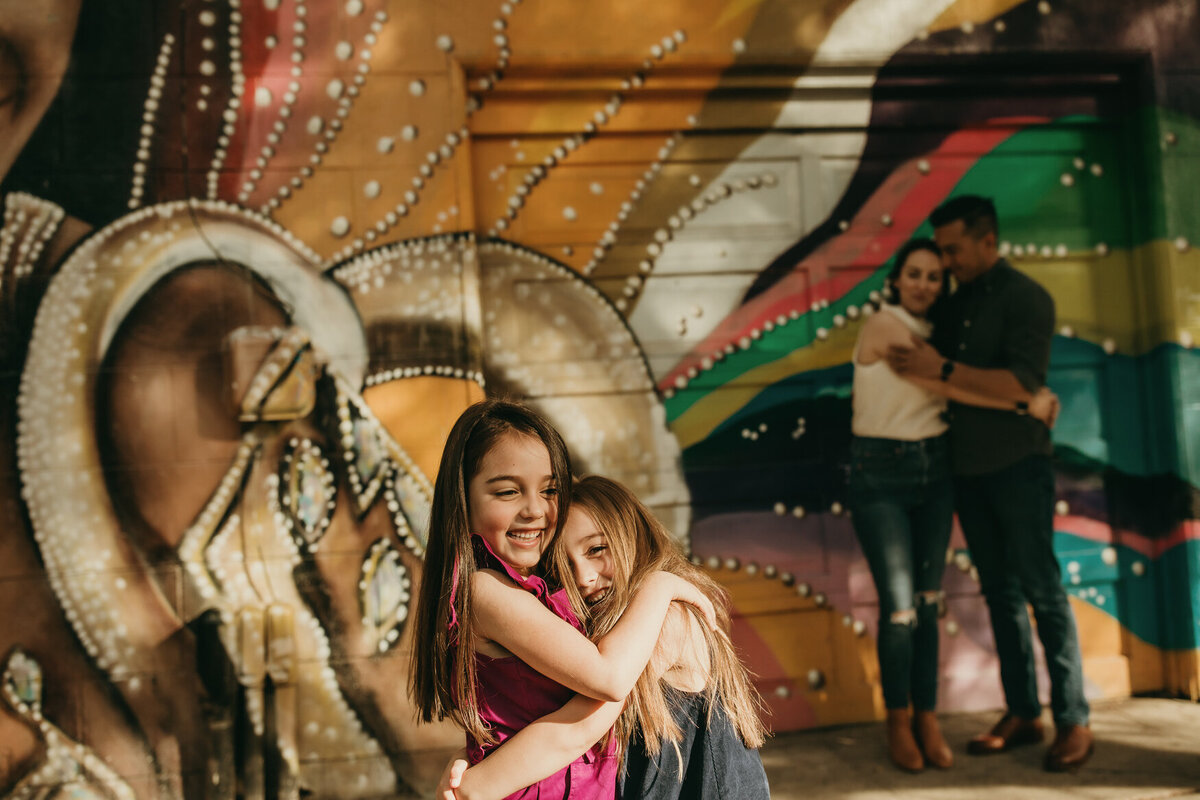 Sisters embrace with colorful mural as backdrop and parents behind them