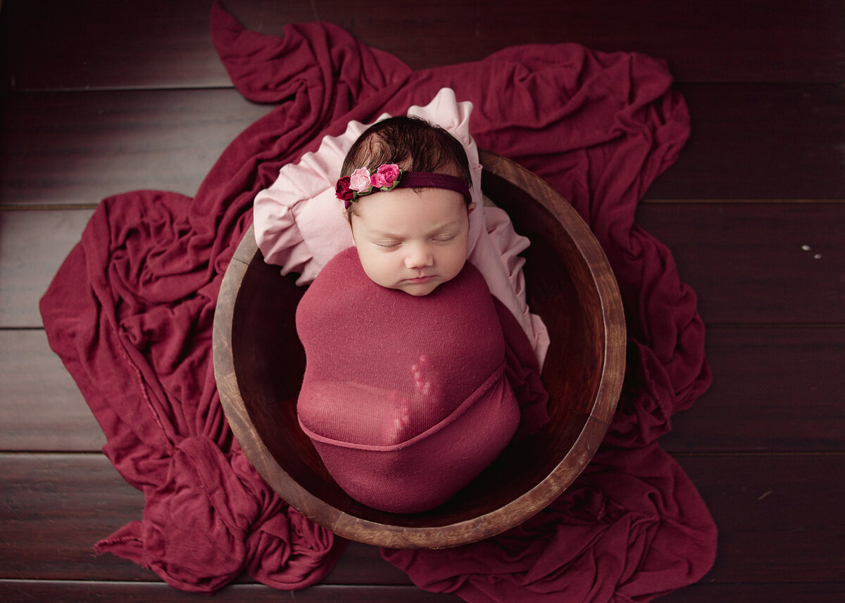 newborn baby girl wrapped in red fabric sleeping in wood bowl during newborn photoshoot in Franklin tennessee photography studio