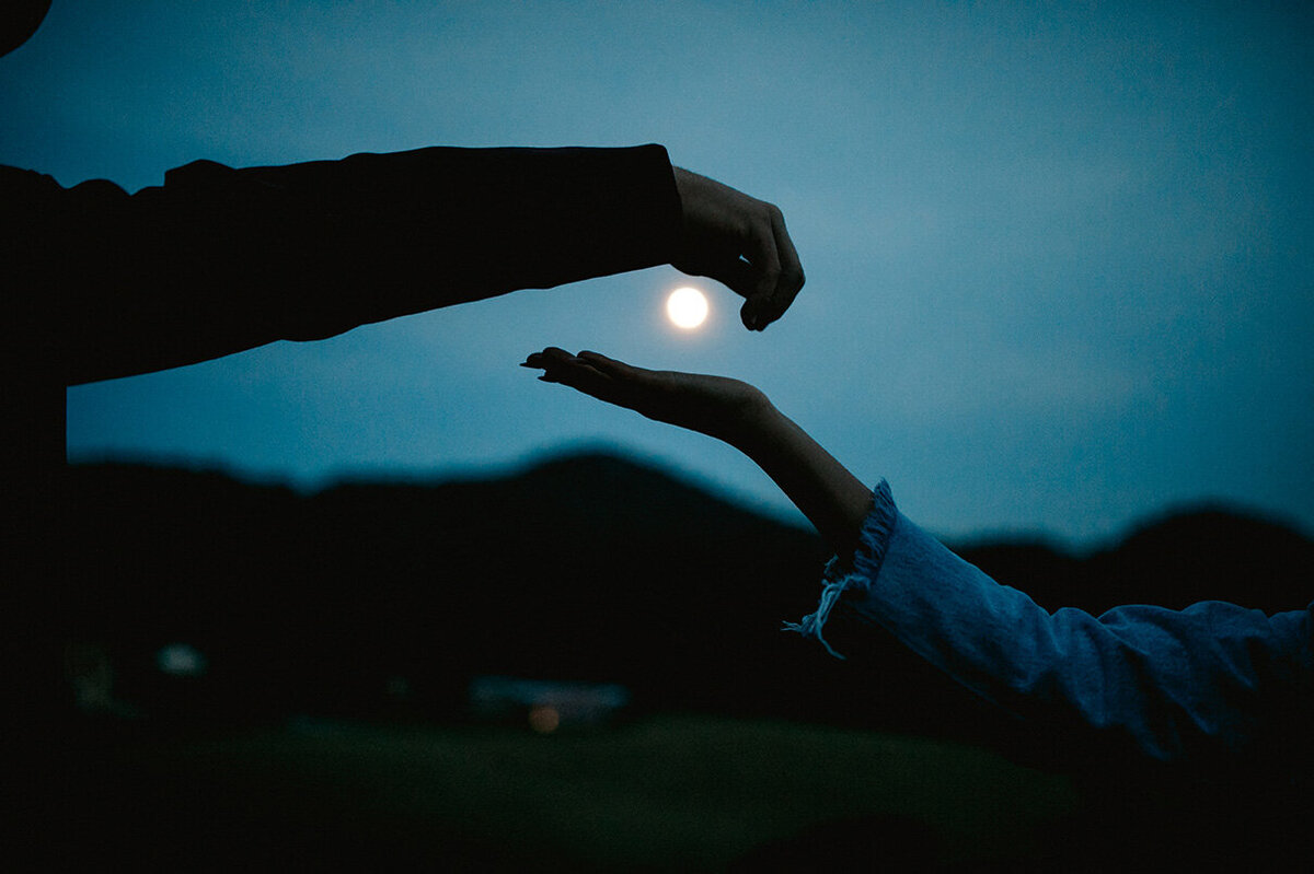 couple poses with the moon in the background in between their hands at dusk