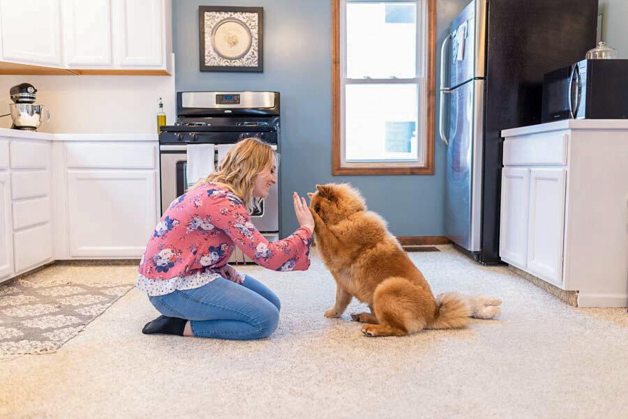 A woman in a floral jacket kneeling in a kitchen, playfully interacting with a fluffy, golden-brown dog.