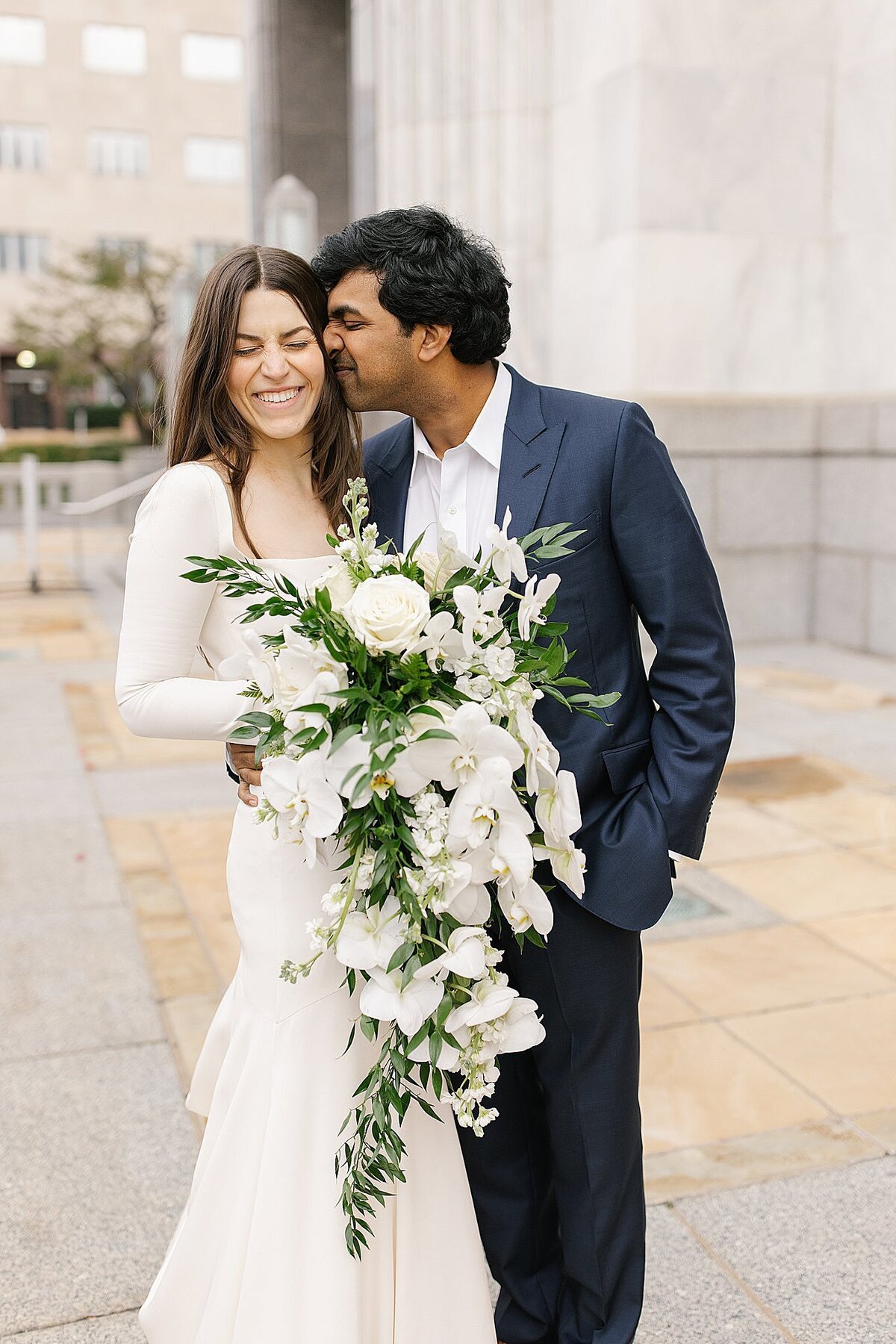 bride in long sleeved fitted white gown laughing  and holding orchid cascade bouquet as groom in dark blue suit kisses her cheek and smiles