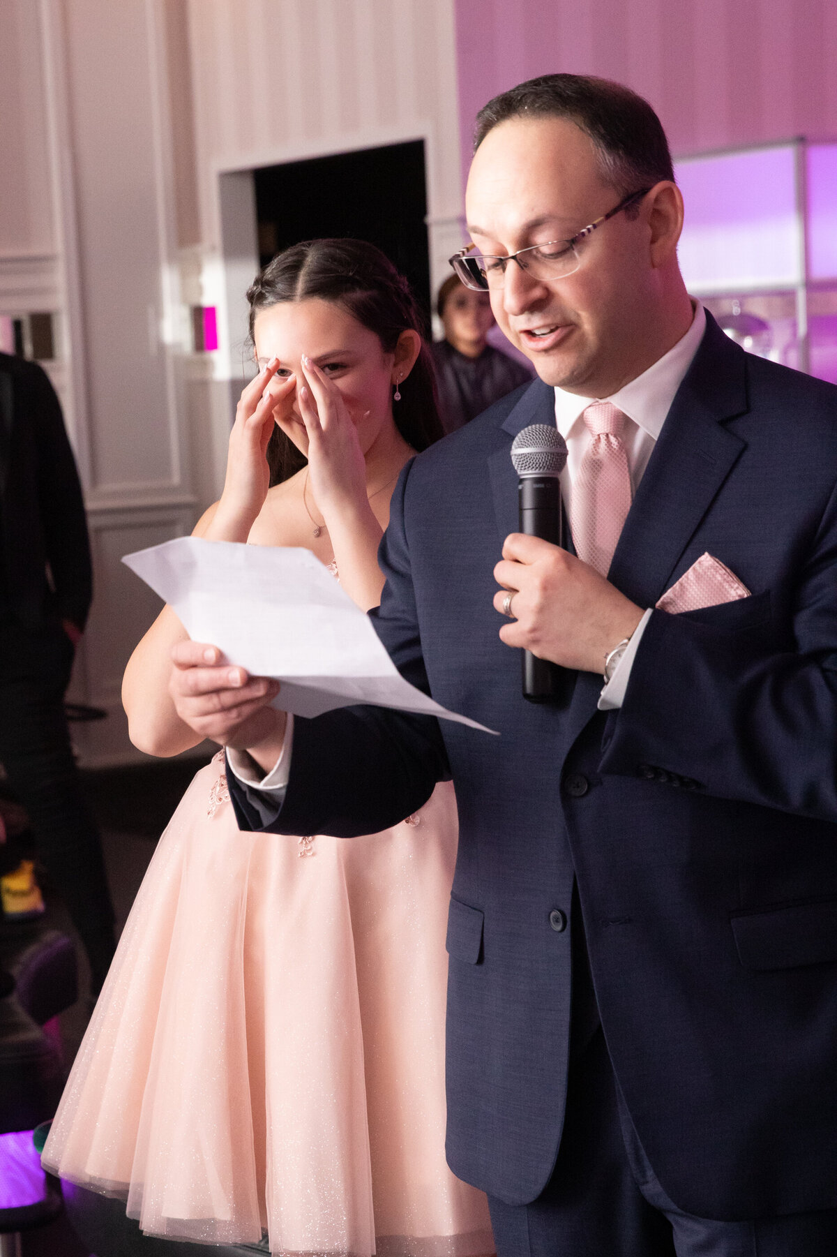 A girl in a pink dress wipes away tears as her father in a blue suit gives a speech at her bat mitzvah Temple Avodah Long Island bar photographer  Milestone Images