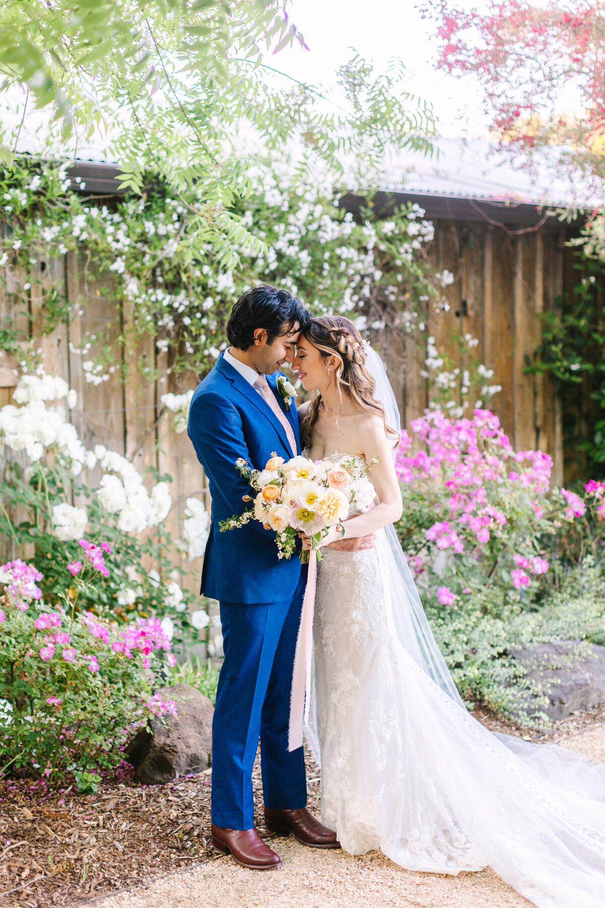 groom in blue suit and bride in white dress with floral bouquet embracing in flower garden.