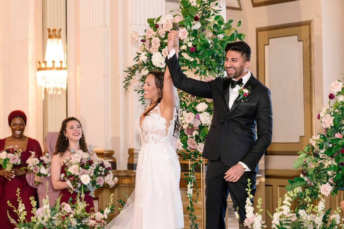 A bride and groom joyfully raise their hands in celebration during their wedding ceremony. The bride wears a white dress, and the groom is in a black suit. Bridesmaids in burgundy dresses hold bouquets, surrounded by elegant floral decorations.