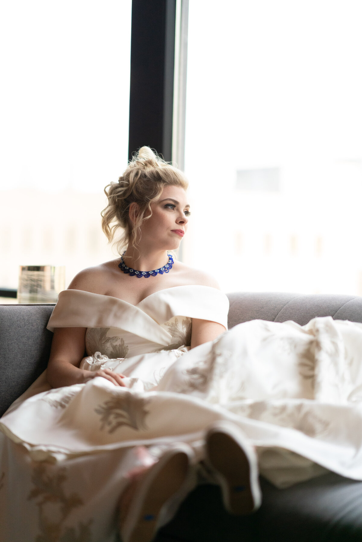 Bride in wedding dress with a blue chocker necklace looks out the window in Saint Paul, Minnesota.