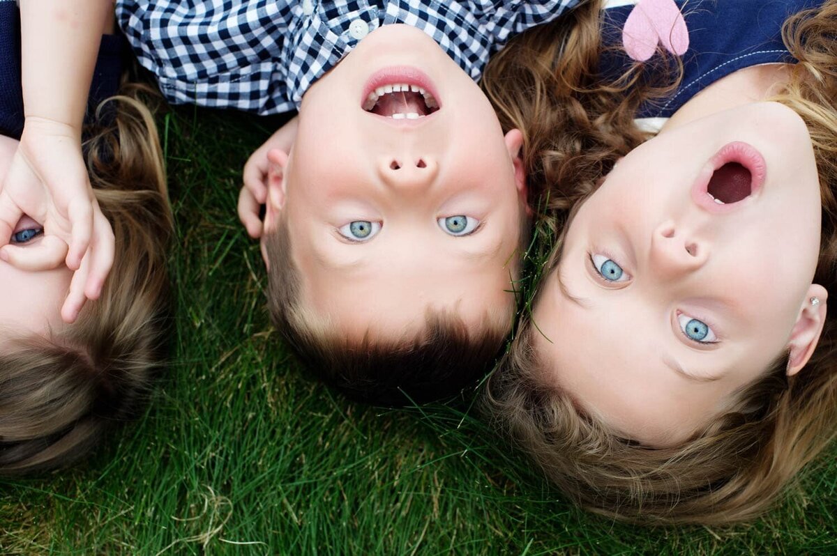 3 blue eyed young siblings laying in the grass together with surprised expressions.