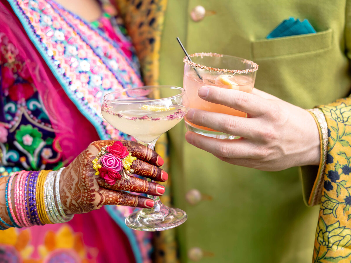 A close-up of a couple holding decorative cocktails. One person, in a colorful outfit with henna-decorated hands and bangles, holds a glass. The other, in a green jacket, holds a different drink. Both drinks have ornate garnishes on the rims.