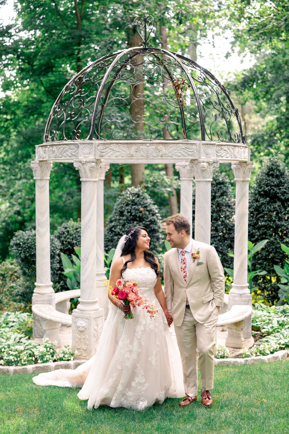 A bride in a lace gown and a groom in a beige suit stand hand in hand, smiling at each other in a garden. They are under an ornate white gazebo, surrounded by greenery and trees.
