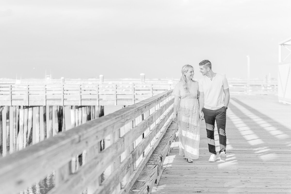 Couple are walking hand-in-hand along a pier in Plymouth, MA for their engagement photoshoot. They are looking at each other to create a candid moment. Captured by New England Wedding Photographer Lia Rose Weddings