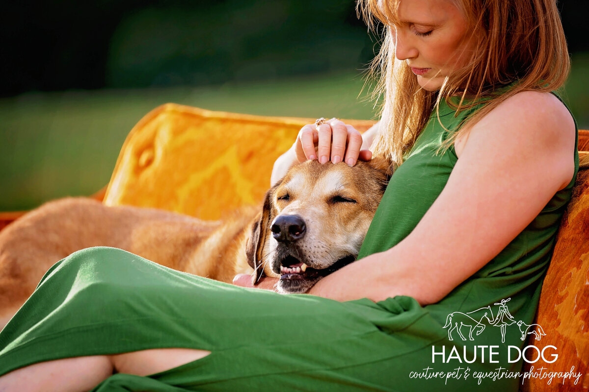 A woman wearing a green dress sitting on an orange couch, affectionately petting a brown dog, who is resting calmly by her side.