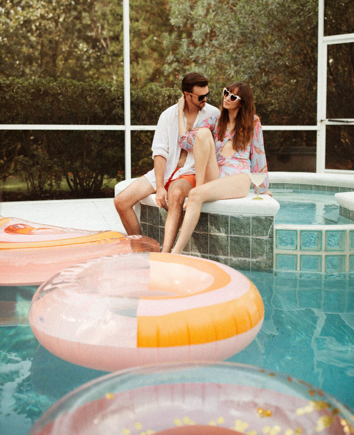 Girl in a retro swimsuit an sunglasses sitting beside a guy in swim trunks and a white button up on the edge of a swimming pool surrounded by floats