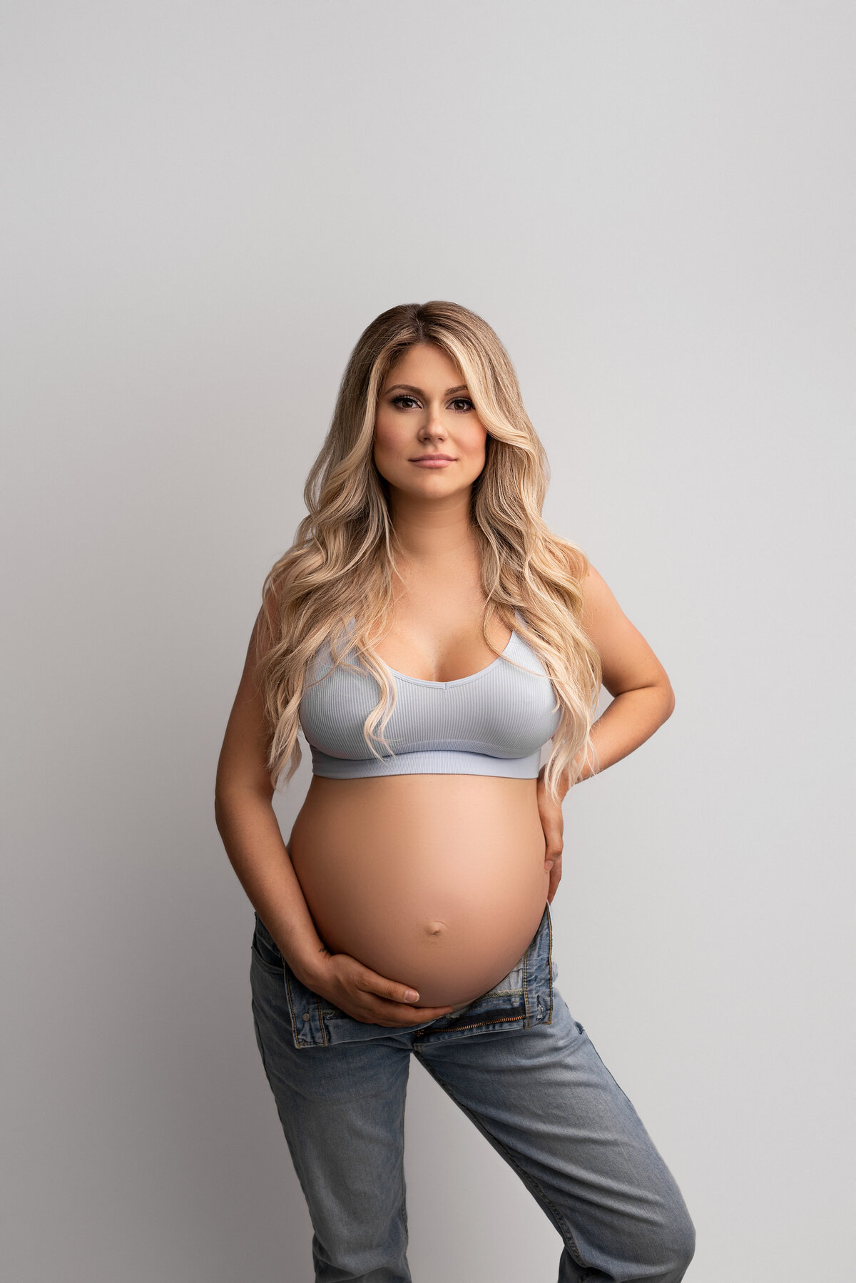 New Jersey's best maternity photographer Katie Marshall captures expectant mom for fine art maternity photos. Woman in unbuttoned jeans and grey sports bra stands facing the camera. One hand is under her bump, the other is at the small of her back. She is looking at the camera with a closed-mouth smile and her long blonde hair trails over her shoulders.