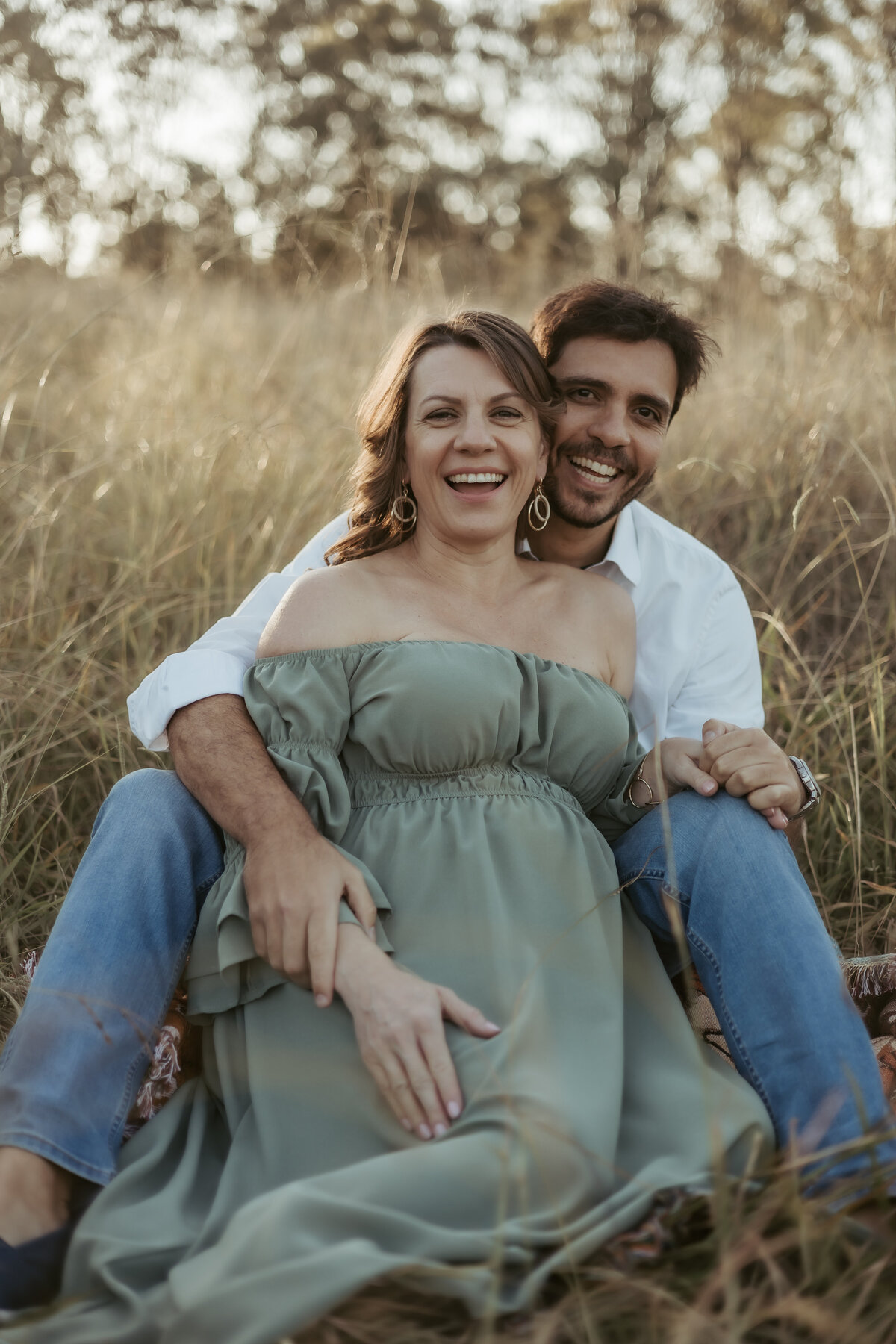 Husband and pregnant wife sitting between his legs and laughing during their comfortable and easy maternity photoshoot in Sydney