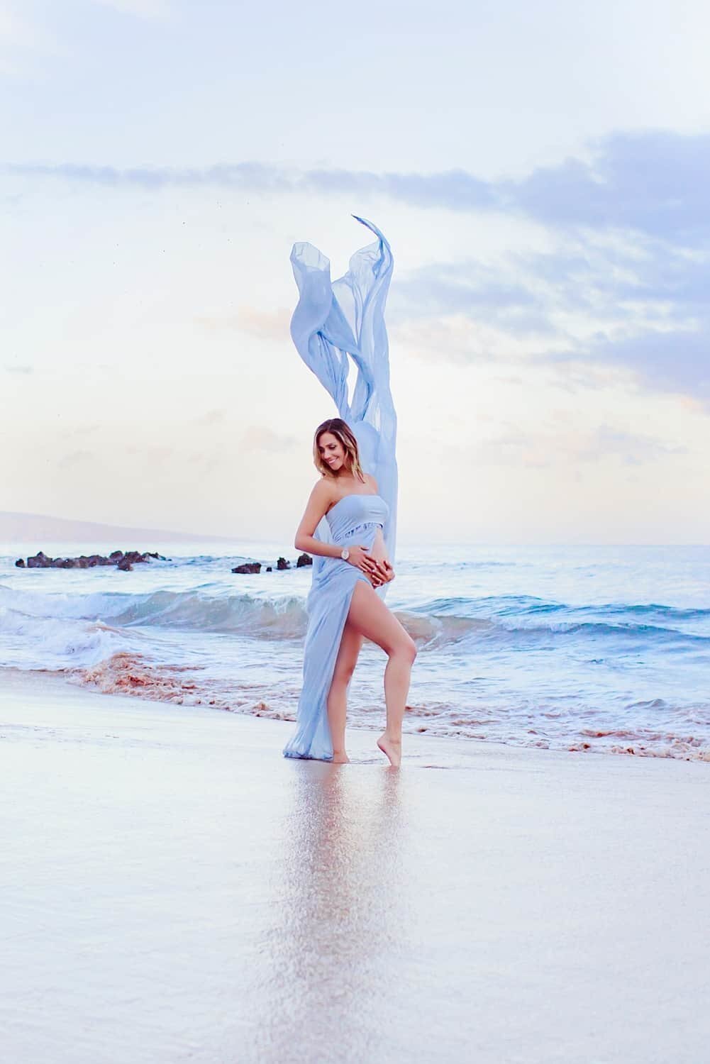 Candid maternity portrait of woman holding belly while her gown flies in the air at sunrise with Love + Water