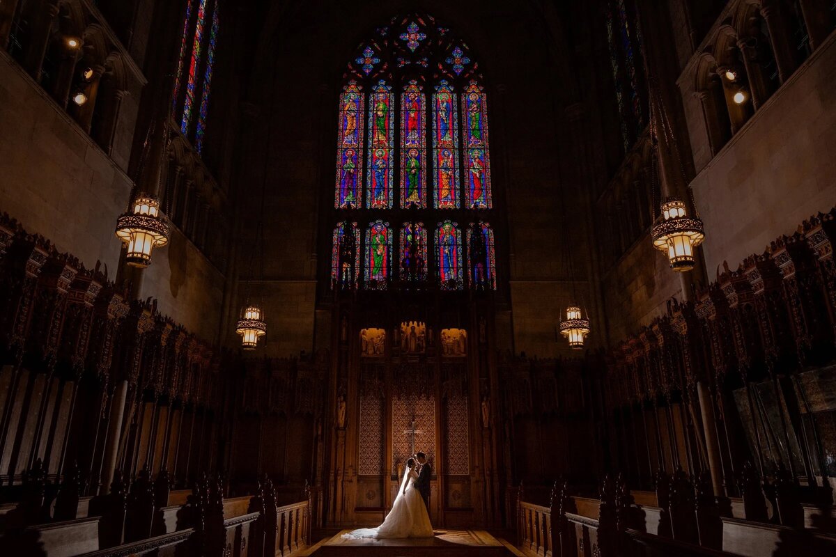 A bride and groom share a moment of intimacy in the stunning gothic interior of Duke Chapel, with vibrant stained glass windows above.