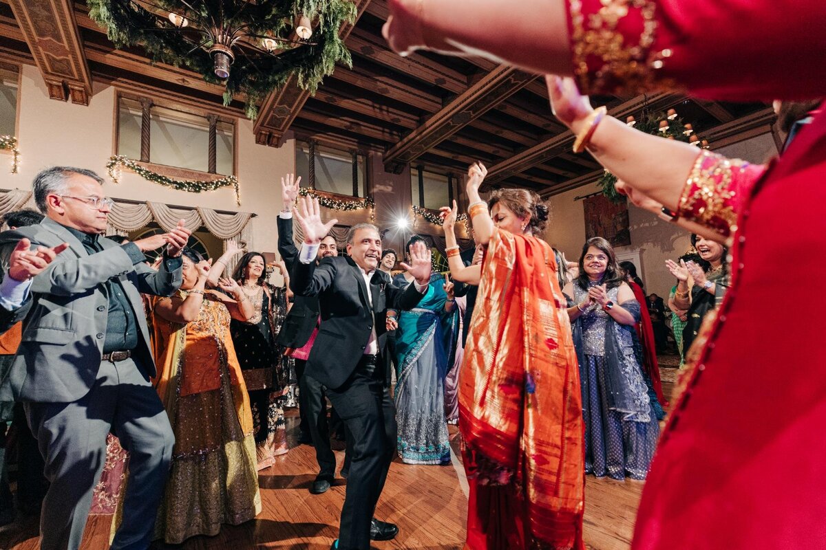 A joyful couple dances at their wedding, surrounded by guests in colorful traditional attire, in a richly decorated hall.