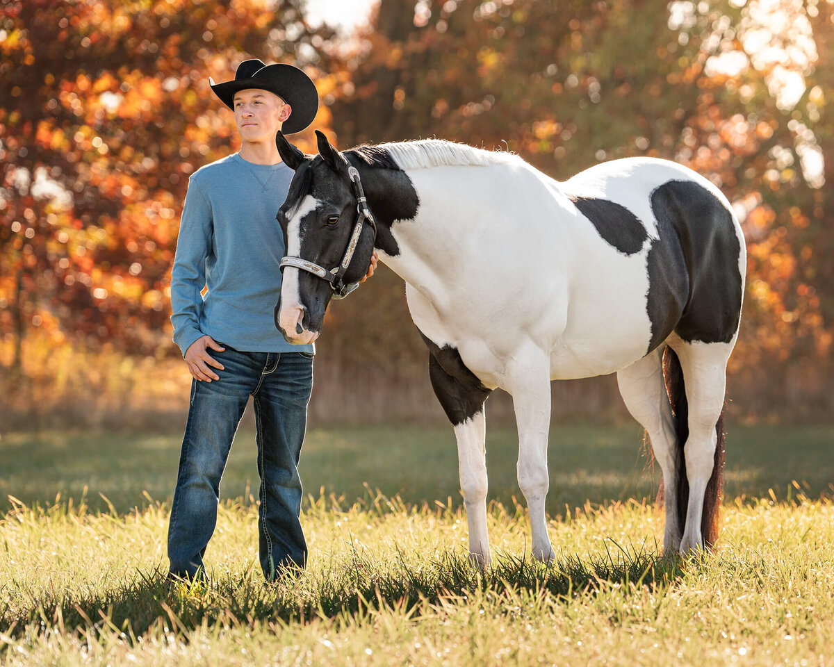 male equestrian wearing a long sleeve blue shirt, jeans, and a black cowboy had standing proudly next to his black and white tobiano Paint horse gelding with beautiful colored fall leaves behind them.