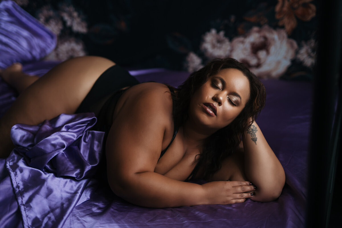 A woman in black lingerie leans her head on her hand on a purple bed