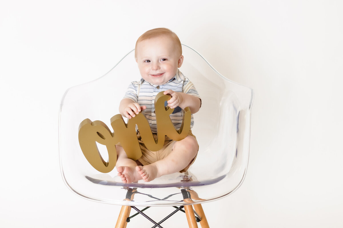 Cake Smash Photographer, a baby holds a carved word "one" on a chair and smiles