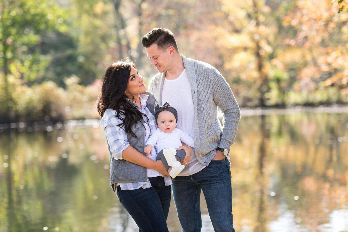 Mother and father, looking at each other while holding their toddler daughter in front of a lake in the fall